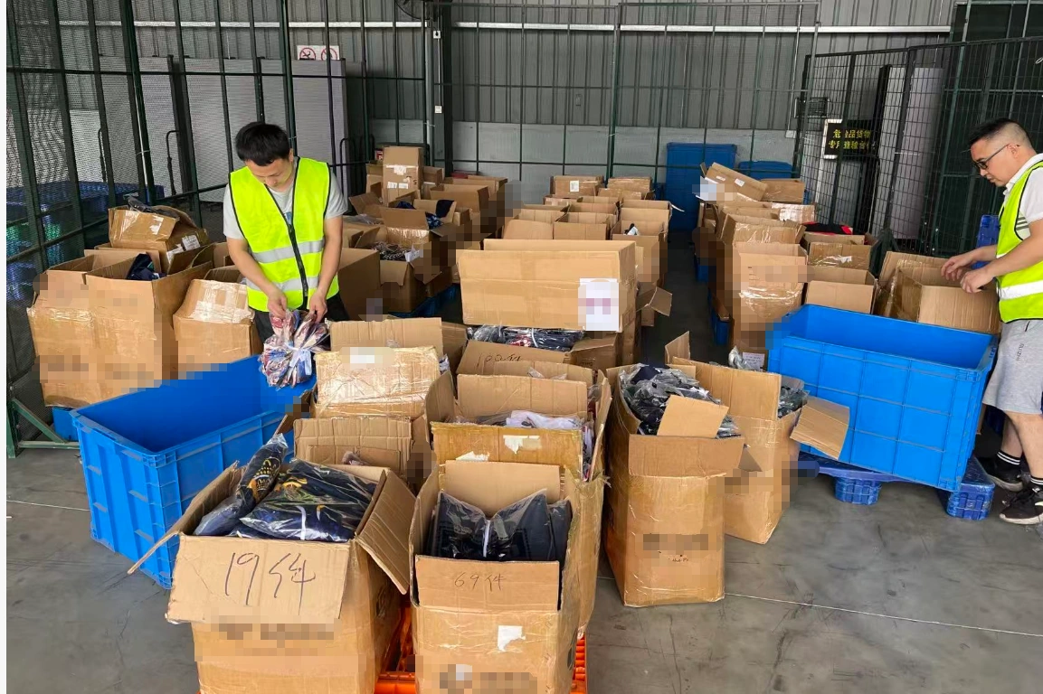 Two workers in high-visibility vests checking packed garments among open cartons and blue bins in an inspection area